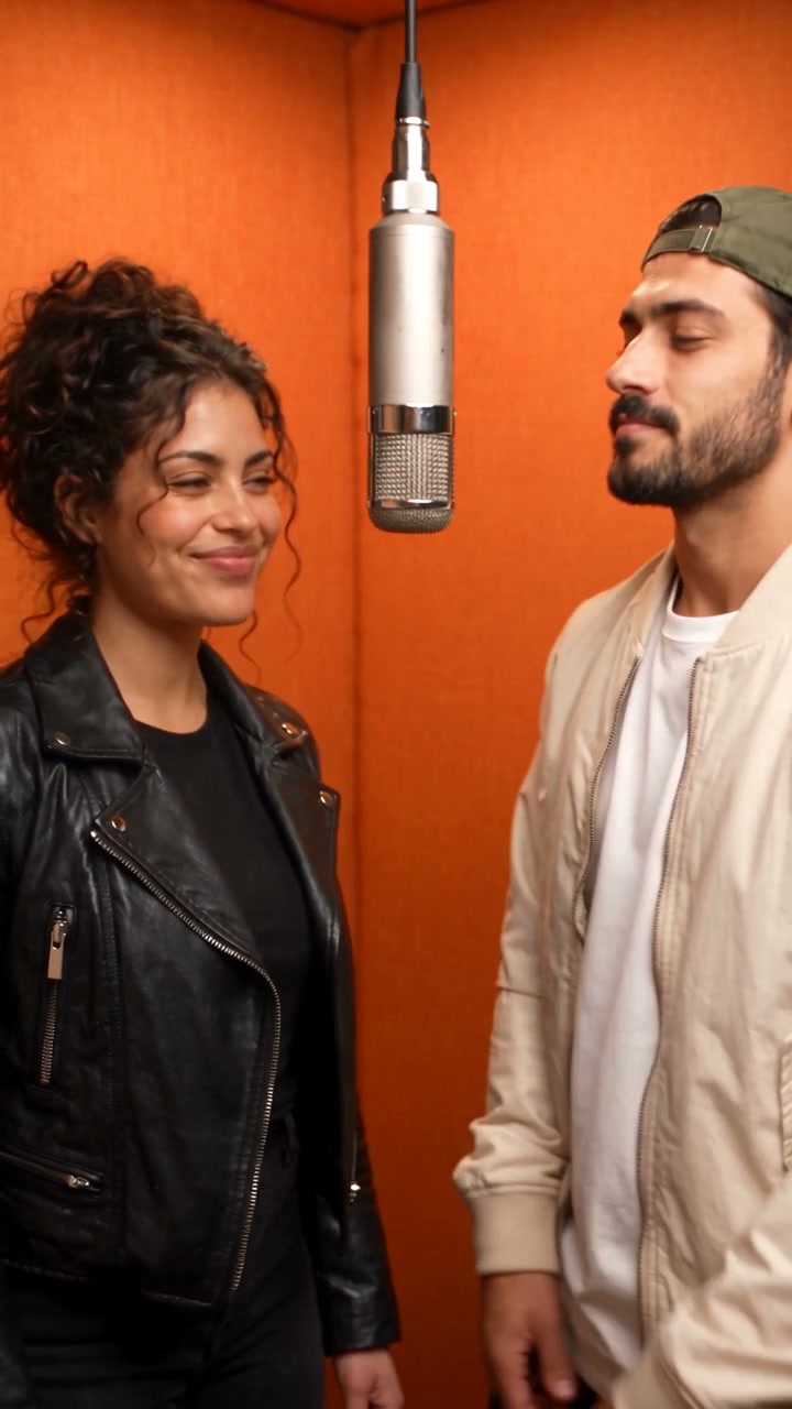 Two fictional adults sharing a silver microphone in a warm orange recording booth